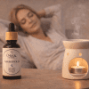 Nightfold oil blend with tealight diffuser Nightfold essential oil blend bottle next to a ceramic tealight oil burner with gentle steam, with a softly blurred person relaxing in the background.
