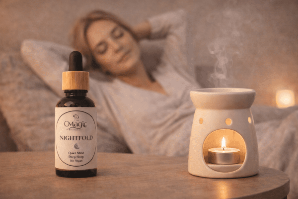 Nightfold oil blend with tealight diffuser Nightfold essential oil blend bottle next to a ceramic tealight oil burner with gentle steam, with a softly blurred person relaxing in the background.